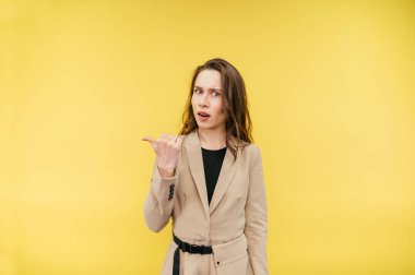 Bej takım elbiseli şaşırmış bir kadın sarı arka planda duruyor, tatminsiz bir yüzle kameraya bakıyor ve bir parmağını yana doğrultuyor..