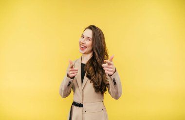 Dişlerinde diş teli olan, bej takım elbiseli neşeli bir kadın sarı arka planda duruyor ve yüzünde bir gülümsemeyle parmaklarını kameraya gösteriyor.