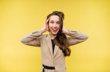 Dişlerinde diş teli ve nedensel giysiler olan neşeli kadın sarı bir arka planda duruyor ve şaşırarak başını kaldırıp kameraya bakıyor ve gülümsüyor..