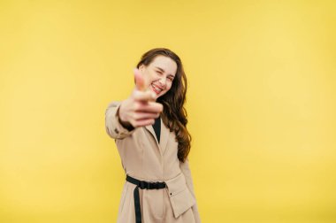Takım elbiseli neşeli kadın parmaklarını gösteriyor ve sarı arka planda neşeli bir yüzle kameraya bakıyor. Kadın kameraya parmağını vuruyor..