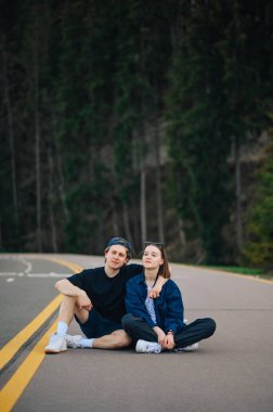 Güzel genç bir çift doğada birlikte vakit geçirir, bir dağ yolunda asfaltta oturur, kozalaklı bir ormanın arka planına yaslanır ve dinlenir..