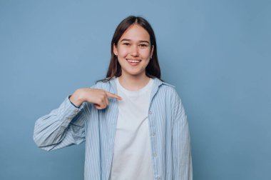 Young woman smiling and pointing at herself against blue background.