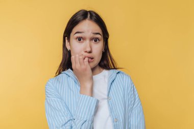 Young woman biting nails, looking anxious against yellow background.