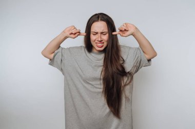 Woman in gray shirt covering ears with fingers, eyes closed.