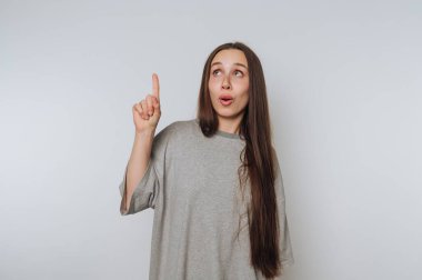 Woman in gray shirt pointing upwards with surprised expression.