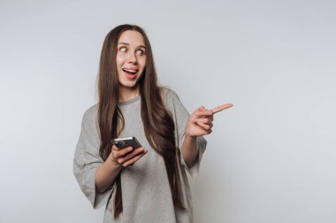 A young woman with long hair holding a smartphone and pointing excitedly to the side against a plain background.