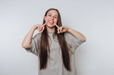 Young woman smiling and pointing at her cheeks against a gray background.