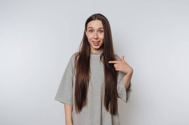 Young woman with long hair pointing at herself, wearing a gray t-shirt, against a plain background.
