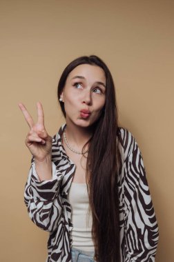 A young woman with long hair making a playful face and peace sign, wearing a zebra-striped shirt against a beige background