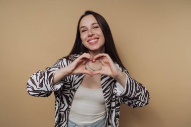 Gülümseyen kadın, elleri bej bir arka planda kalp şekli yaratıyor..