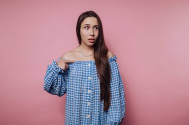A young woman with long hair wearing a blue and white checkered off-shoulder top, looking surprised and pointing at herself against a pink background
