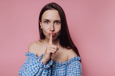 Woman in blue checkered off-shoulder top making a shushing gesture against a pink background.
