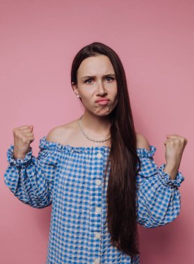 Woman with long hair in a blue checkered dress showing frustration against a pink background.