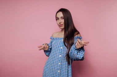 A young woman in a blue gingham off-shoulder dress posing playfully against a pink background