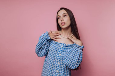 A young woman in a blue and white checkered off-shoulder top posing against a pink background, showcasing a necklace