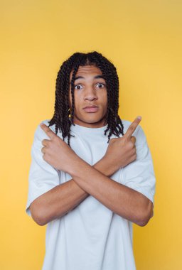 Young man with dreadlocks crossing arms and pointing fingers on yellow background.