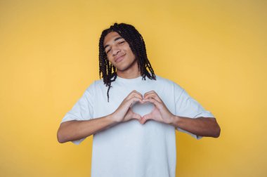 Young man with braided hair making a heart shape with hands against a yellow background.