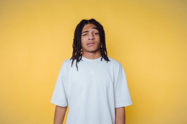 A young person with braided hair, wearing a light grey t-shirt, standing against a bright yellow background, displaying a confused expression