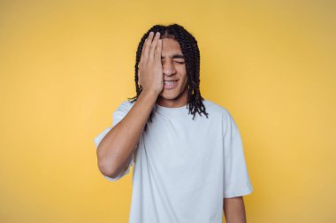 Young person with dreadlocks covering face with hand, yellow background.