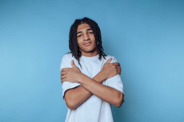 Young man with braided hair hugging himself against a blue background.