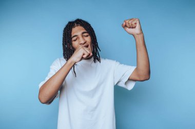 A young man with braided hair wearing a white t-shirt, dancing joyfully against a blue background