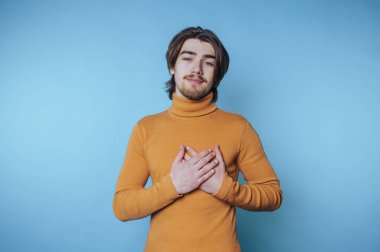 Young man in mustard turtleneck with hands on chest against blue background.