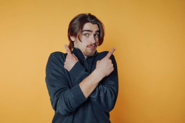 Young man with long hair and beard, wearing a dark sweater, standing against a yellow background, crossing arms and pointing in opposite directions with both hands.