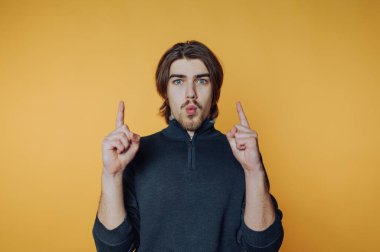 Young man with surprised expression pointing upwards against yellow background.