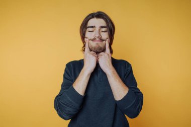 A young man with long hair and a beard, smiling and making a playful expression against a bright yellow background