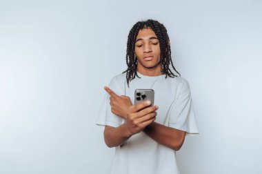 Young man with braided hair pointing at smartphone, neutral background.