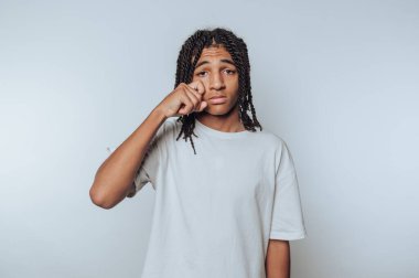 A young person with braided hair wiping a tear from their eye against a plain background.