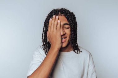Young man with dreadlocks covering one eye with his hand, smiling.