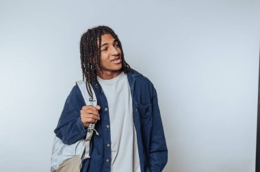 Young man with braided hair holding a backpack, wearing a denim jacket and white t-shirt, looking to the side against a plain background.