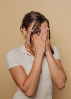 A woman in a beige outfit covers her face with her hands against a beige background.