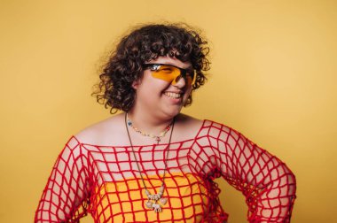 Smiling person with curly hair wearing yellow glasses and a red net top against a yellow background.