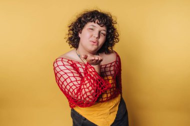 Person blowing a kiss, wearing a red net top over a yellow shirt, against a yellow background.