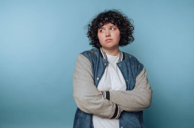 A person with curly hair wearing a varsity jacket and white shirt, looking to the side against a blue background.