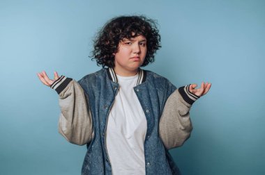 Person with curly hair in a denim jacket shrugging against blue background.