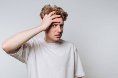 Young man in a white t-shirt looking confused, hand on forehead.