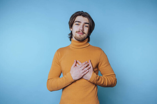 Young man in mustard turtleneck with hands on chest against blue background.