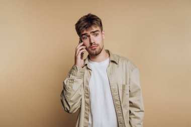 Endişeli bir ifadeyle genç bir adamın portresi telefonda iletişim kuruyor, bej arka planda endişe ve tefekkür duygularını gösteriyor.