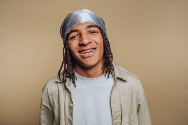 Genç ve neşeli bir adamın portresi geniş bir gülümsemeyle bej arka planda gümüş bir başlık takarak diş tellerini gösteriyor.