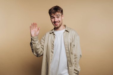 Genç gülümseyen adamın portresi kameraya neşeyle el sallıyor, bej arka planda arkadaşlığı somutlaştırıyor.