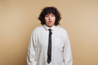Kıvırcık saçlı, şaşırmış genç bir kadının portresi, bej arka planda yakalı gömlek ve kravat takarken şoka giriyor.