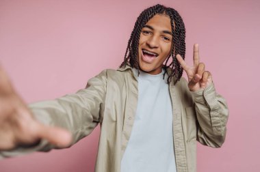 Genç ve neşeli bir adamın portresi barış işareti veriyor, pembe arka planda pozitiflik ve neşe yayıyor