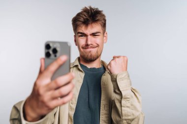 Heyecanlı genç adamın portresi, akıllı telefonunu beyaz arka planda tutarken muzaffer bir ifade sergiliyor.