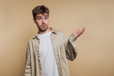 Genç bir adamın portresi, bej arka planda açık bir avuç içi jesti ile kafa karışıklığı göstererek rahat bir poz verir.