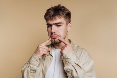 Genç bir adamın portresi hüzünlü bir ifade sergiliyor. Kırılganlığını vurgulayarak bej arka plandaki çatık kaşlarına işaret ediyor.