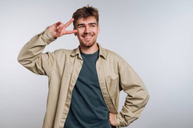 Neşeli genç adamın portresi gülümserken bir barış işareti gösteriyor, beyaz arka planda pozitiflik ve dostluk yayıyor.