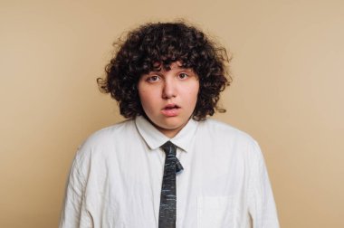 Kıvırcık saçlı genç bir kadının portresi bej arka planda şaşkınlık dolu bir bakış sergiliyor.